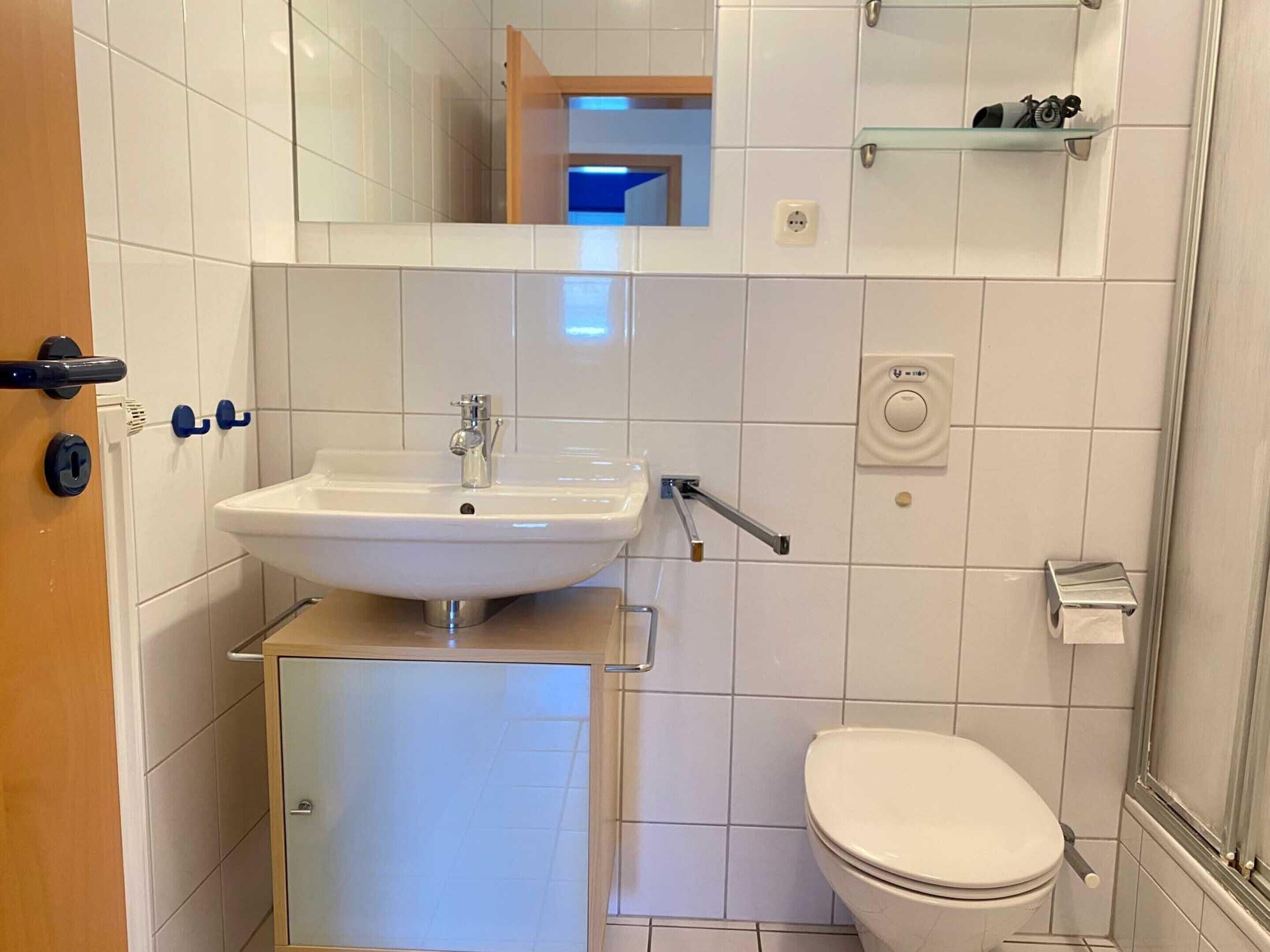 Un baño con lavamanos blanco, inodoro, espejo y estante con artículos de aseo.