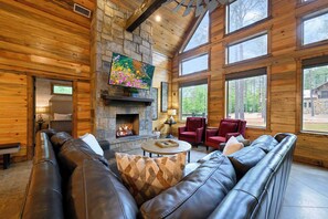 A log cabin interior with a stone fireplace, leather sofas, and large windows. - Sleeps 21 • 4 King Ensuites • Game Room, Hot Tub & Outdoor Fun (Broken Bow)