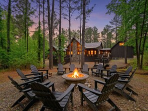 A lodge with a fire pit surrounded by Adirondack chairs, set in a forested area with a stone building in the background. - Sleeps 21 • 4 King Ensuites • Game Room, Hot Tub & Outdoor Fun (Broken Bow)