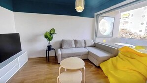 Una sala de estar moderna con un televisor de pantalla plana, un sofá gris, una mesita redonda pequeña, una planta en maceta y un ventanal con vistas a edificios.