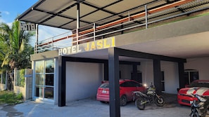Un hotel llamado 'HOTEL JASLI' con estacionamiento techado, un mostrador de recepción y una motocicleta estacionada afuera.