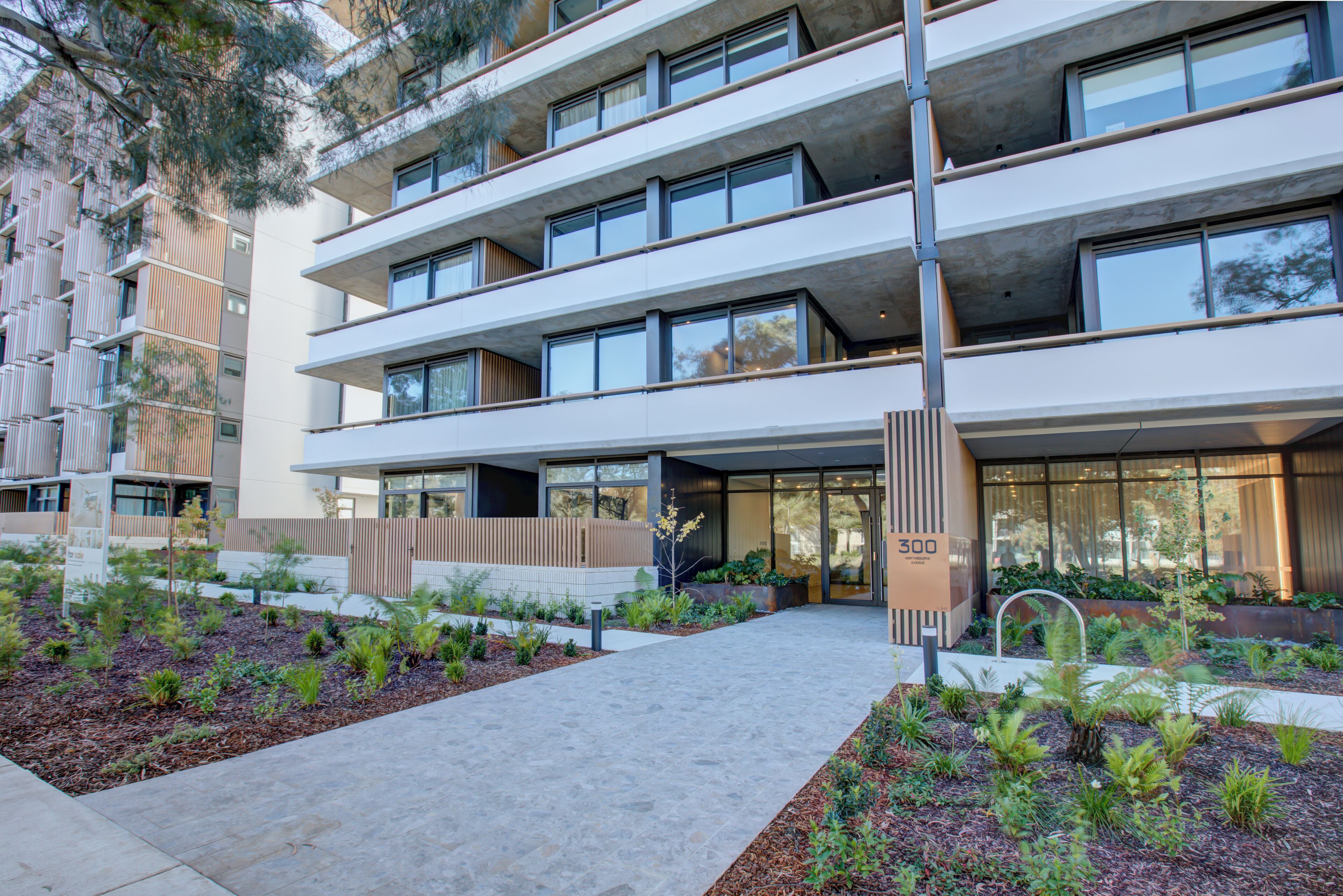 A multi-story building with balconies, a central entrance, and landscaped areas with plants.