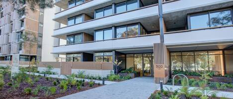 A multi-story building with balconies, a central entrance, and landscaped areas with plants.