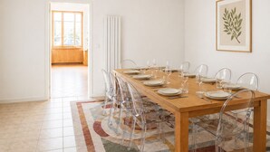 Uma sala de jantar com mesa de madeira posta para seis, cadeiras transparentes, radiador e planta emoldurada na parede.