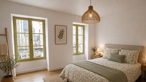 Quarto com uma cama grande, duas janelas com molduras verdes, um candeeiro pendente e um vaso de planta.