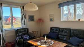 A living room with a leather sofa, a wooden coffee table with a candle, a black chair, a red house visible through the window, and a lamp. -  The Old Farmhouse (Hammarstrand)