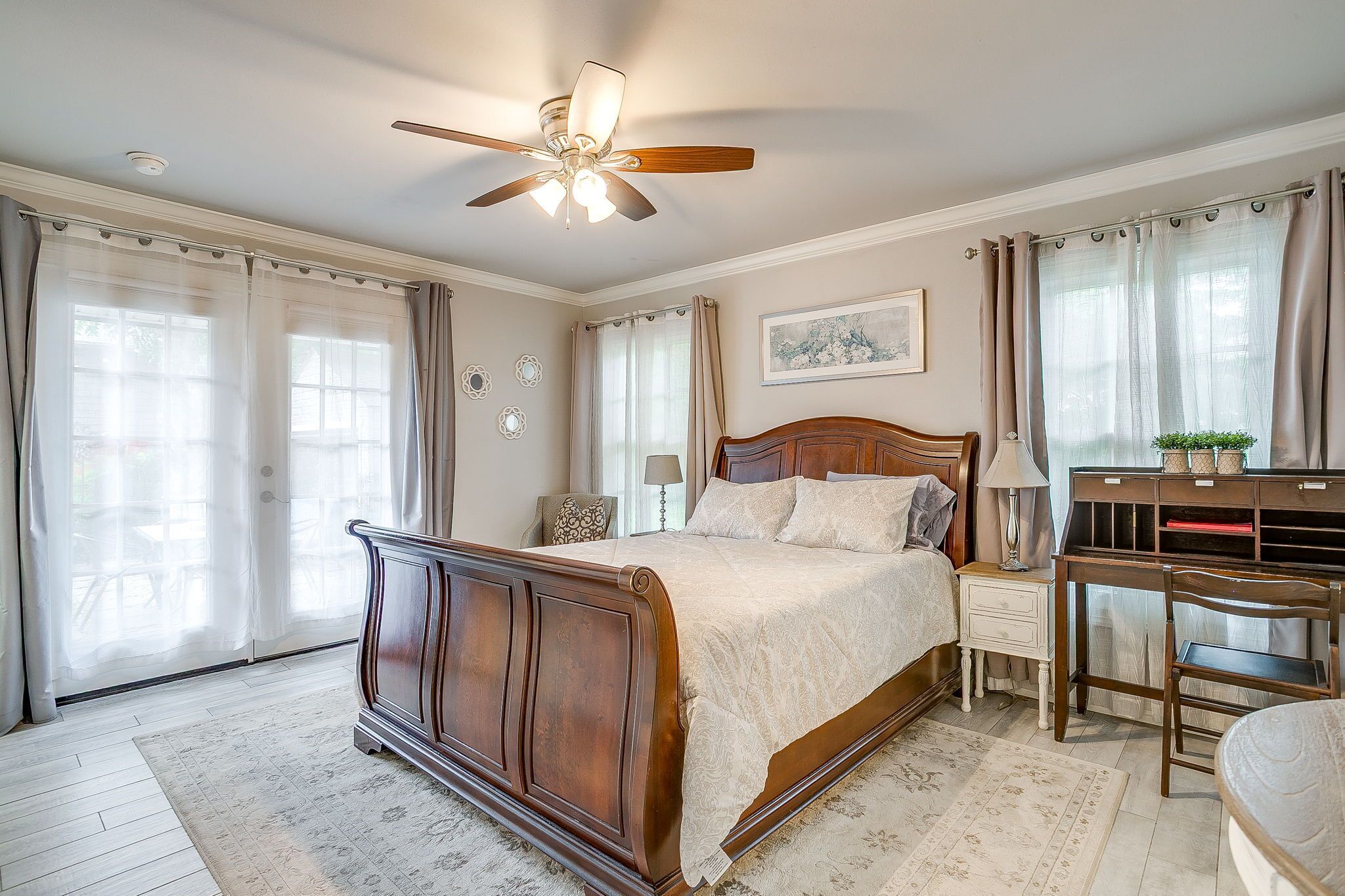 A bedroom with a wooden bed, a ceiling fan, a nightstand, a desk, and a window with curtains.