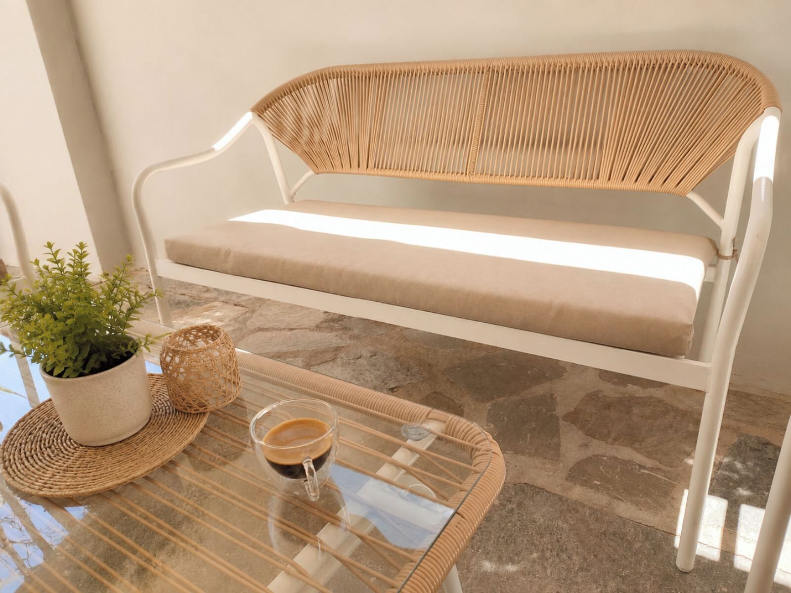Eine weiße Metallbank mit einem gewobenen Sitz und Rücken, ein beiges Kissen, ein Glastisch mit Untersetzer aus Bicolin, ein Blumentopf und eine Kaffeetasse.