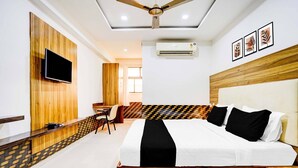 A hotel room with a bed, a desk, a chair, a TV mounted on a wooden panel, an air conditioner, and framed artwork on the wall.