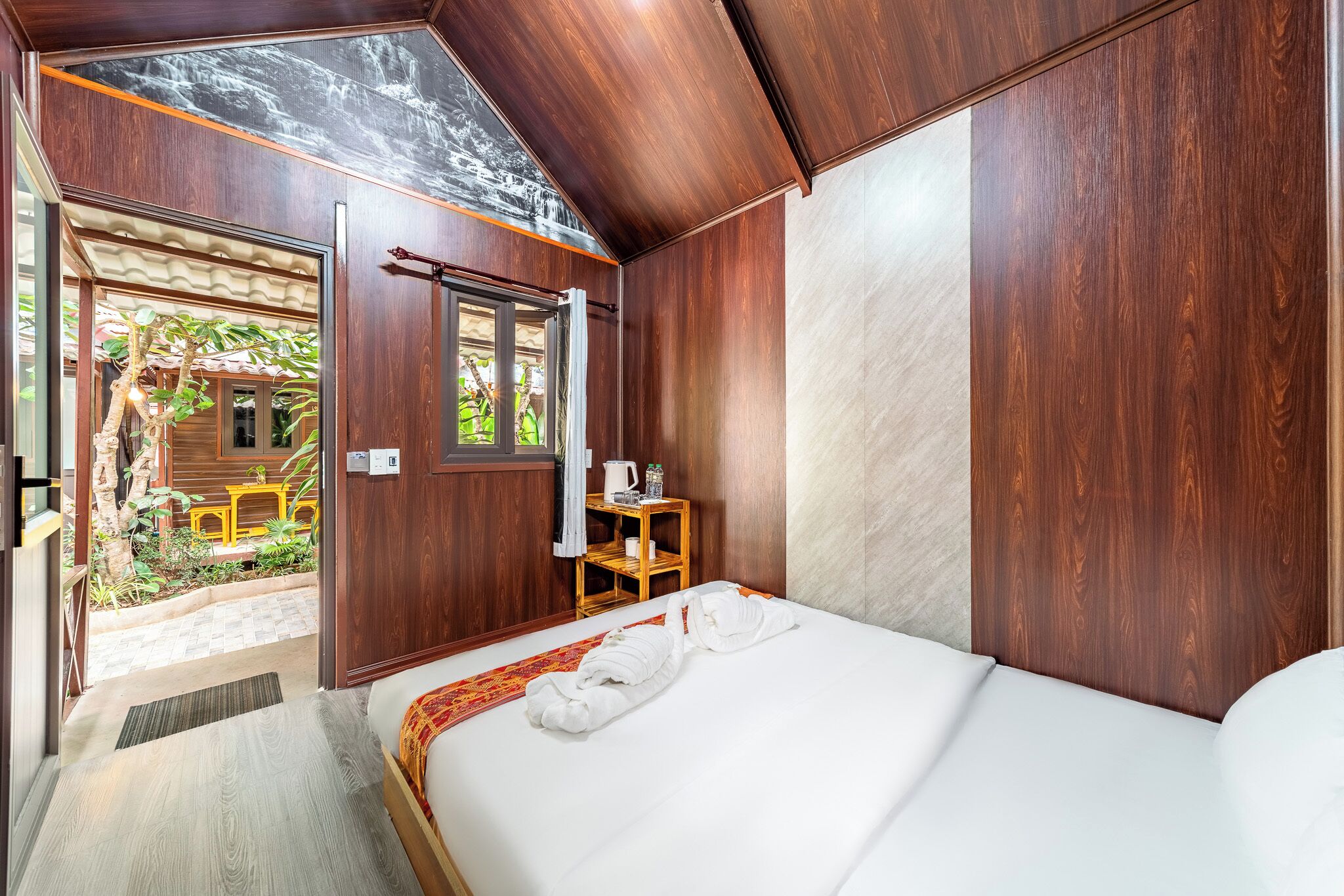 A room with a bed, a small table with a kettle, and a window with a view of greenery.
