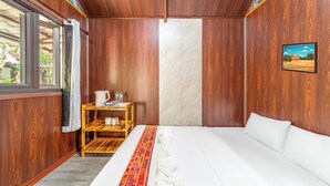 Habitación de hotel con cama, una pequeña mesa de madera con una tetera y botellas, una ventana con cortinas y un cuadro en la pared.