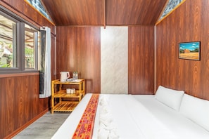 Une chambre d’hôtel avec un lit, une petite table en bois sur laquelle se trouvent une bouilloire et des bouteilles, une fenêtre avec des rideaux et un tableau encadré accroché au mur.