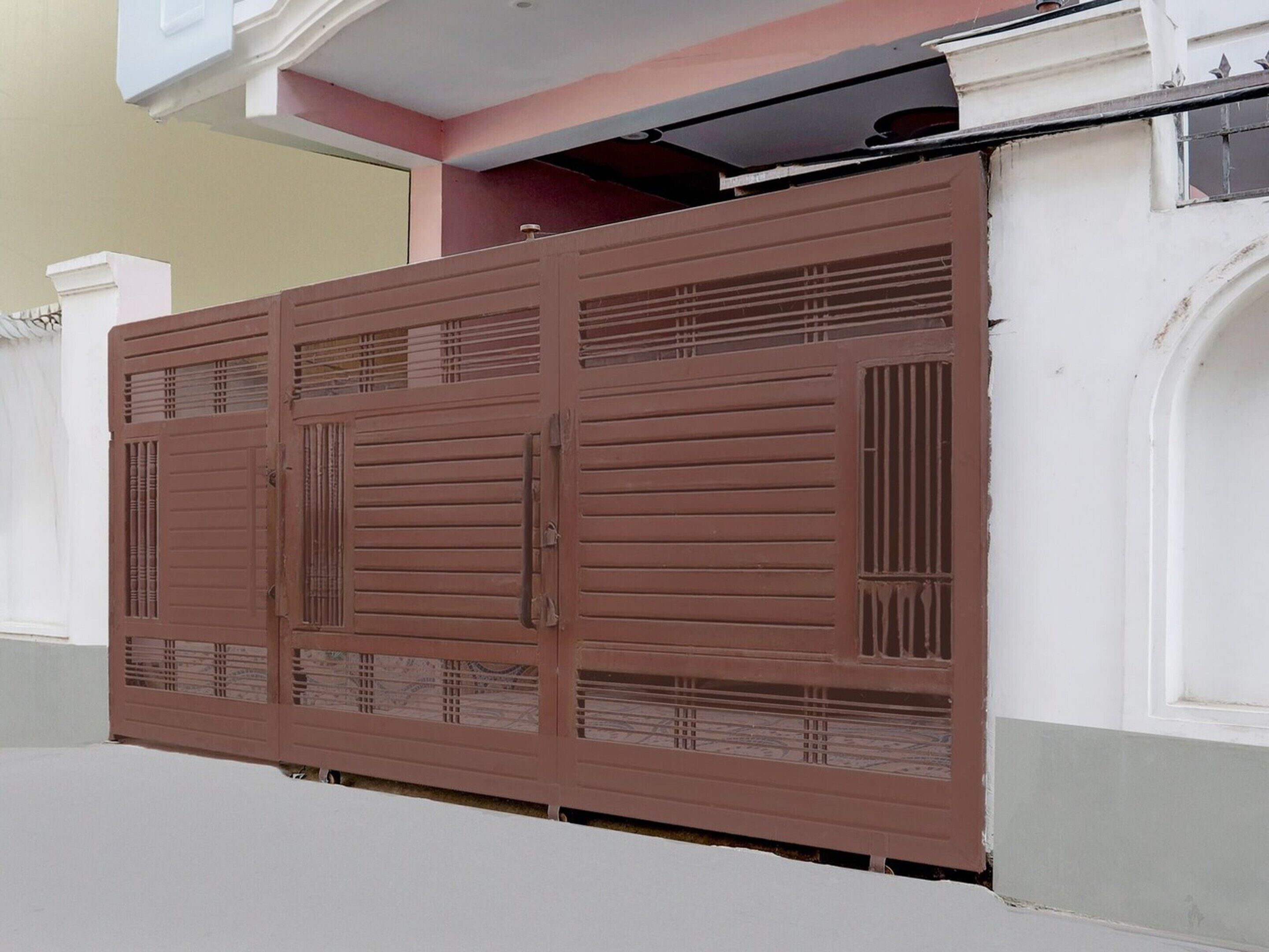 Ein braunes Metallgitter mit waagerechten Streben und einem Riegel, angebracht an einem Gebäude mit weißer Fassade und rosa Dach.
