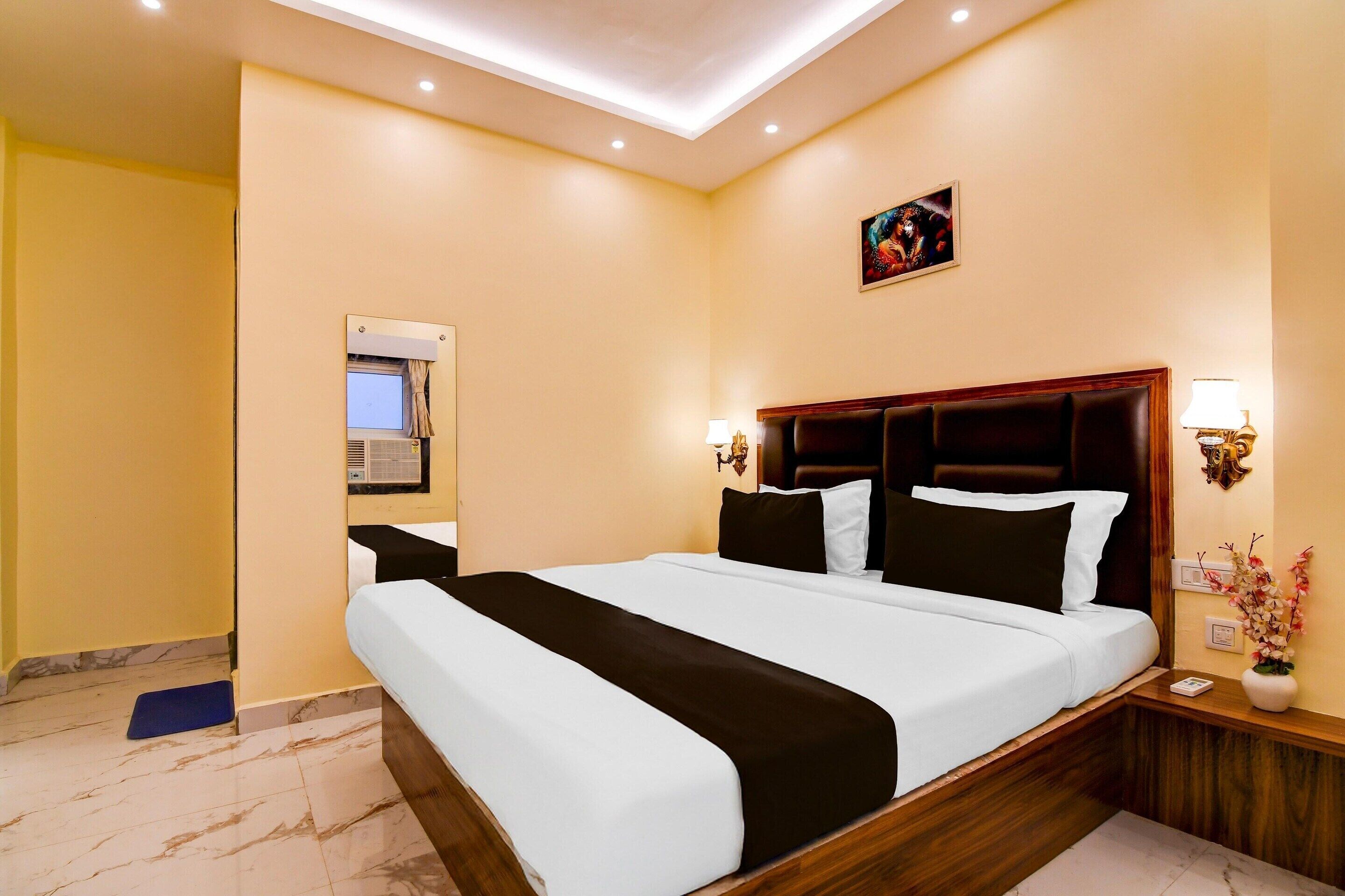 A hotel room with a large bed, a bedside table with a lamp, a wall-mounted air conditioning unit, and a small picture on the wall.