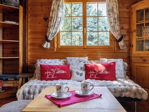 Interior acogedor de una cabaña con una cama de madera, dos almohadas, una mesa con dos tazas y una ventana con cortinas.
