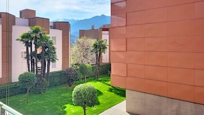 Un edificio moderno con facciata rossa, un giardino curato con palme e una vista chiara sulle montagne lontane.