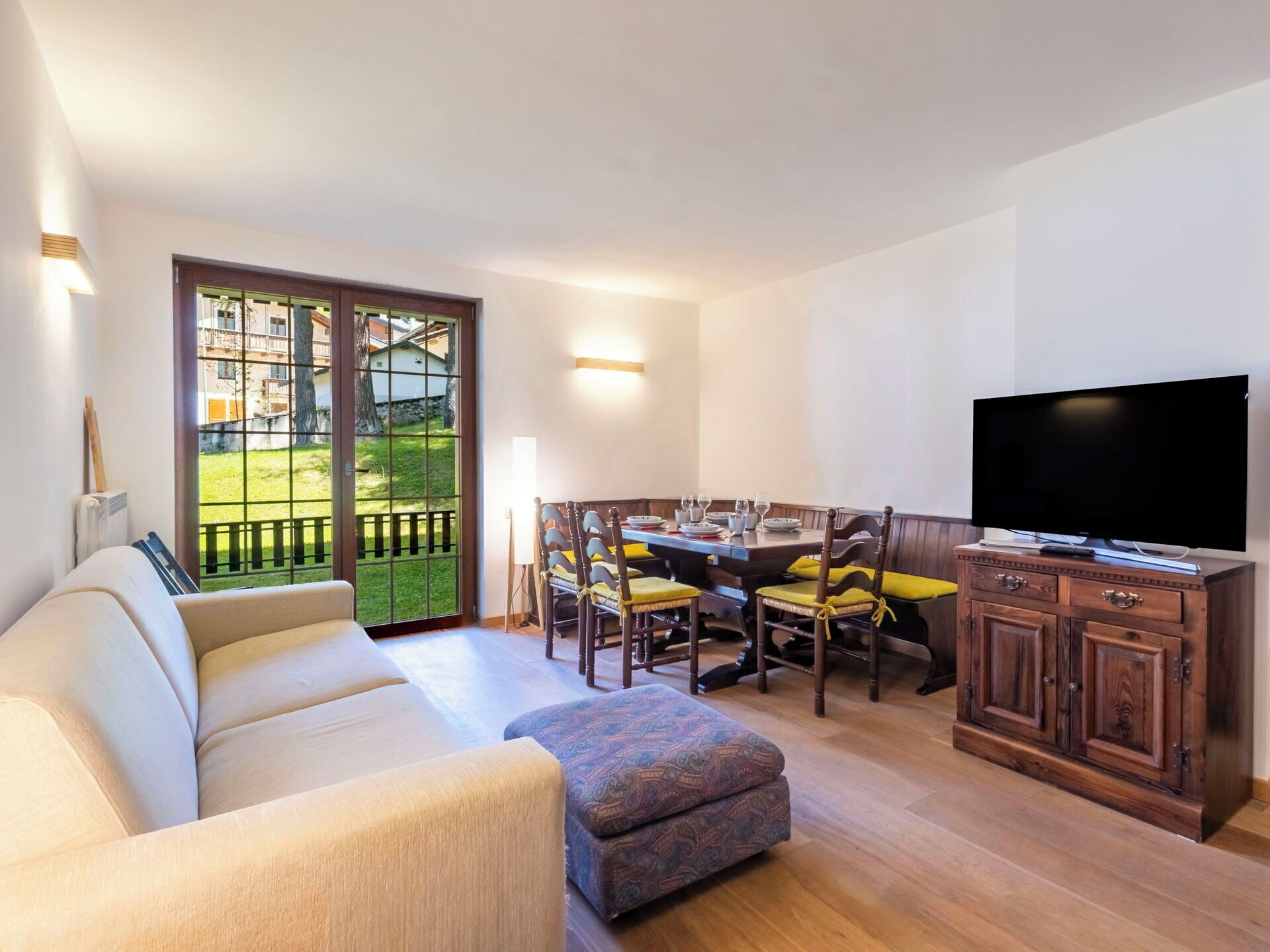 A living room with a sofa, ottoman, dining table with chairs, a TV, and a view through a glass door.