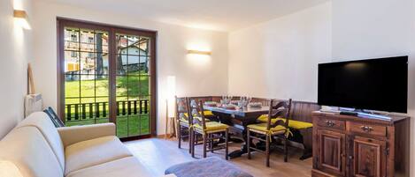A living room with a sofa, ottoman, dining table with chairs, a TV, and a view through a glass door.
