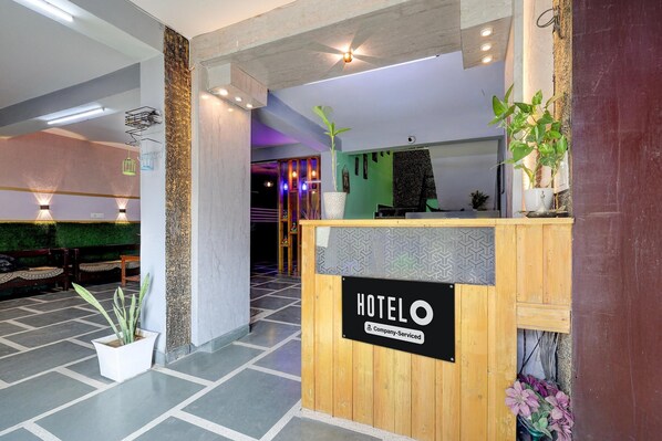 A hotel reception desk with a sign, a potted plant, and a seating area in the background.