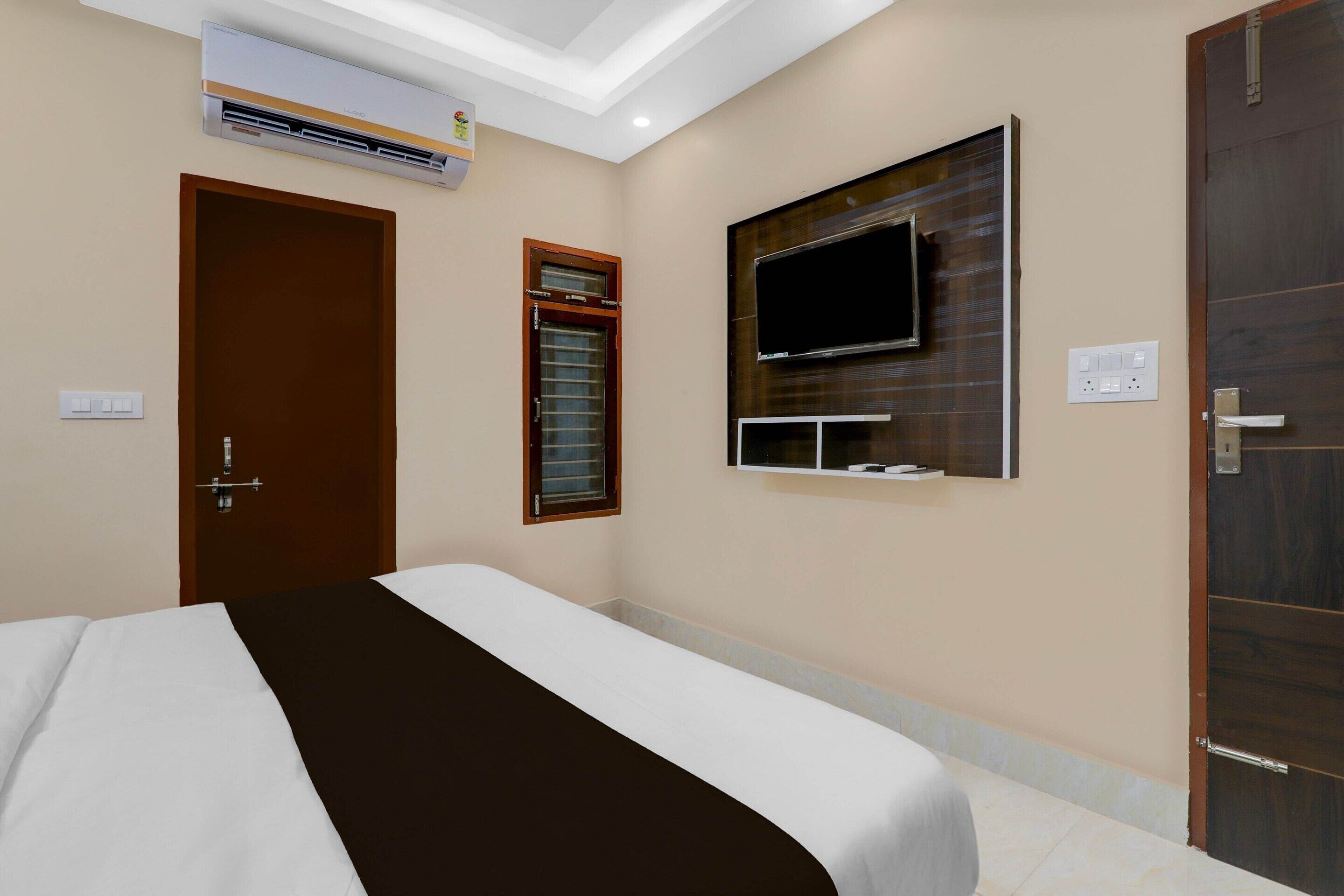 A hotel room with a bed, an air conditioning unit, a television mounted on the wall, a door, and a window with blinds.