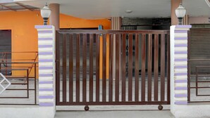 A gated entrance with a metal gate, a concrete pathway, and a building with orange walls and a white roof. - Deluxe Pushp Vatika (Varanasi)