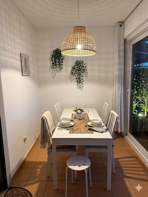Un coin repas avec une table blanche dressée pour quatre personnes, un luminaire suspendu et des plantes en pot.