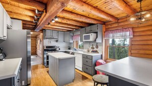 A kitchen with wooden beams, a central island, grey cabinets, a microwave, and a window with a checkered valance. - Hilltop Log Cabin Retreat with Hot Tub & Direct Trail Access Near Lead (Lead)