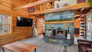 A log cabin interior with a stone fireplace, wooden walls, a television, and a leather sofa. - Hilltop Log Cabin Retreat with Hot Tub & Direct Trail Access Near Lead (Lead)