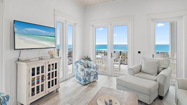 Un salón amplio con un televisor grande, un sofá blanco, un sillón estampado y un mueble con puertas de cristal.