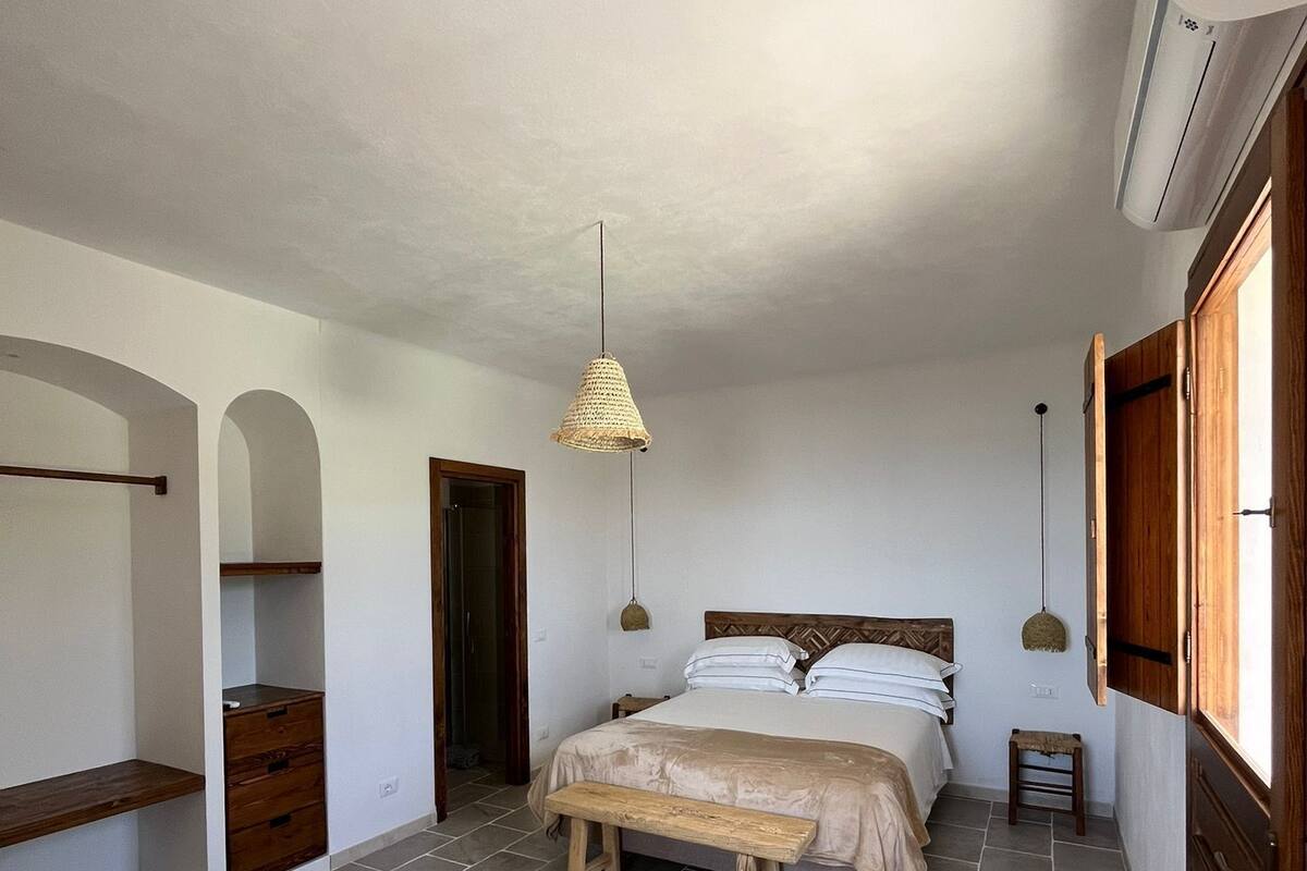 A bedroom with a bed, a bench, a hanging light, and an air conditioning unit.