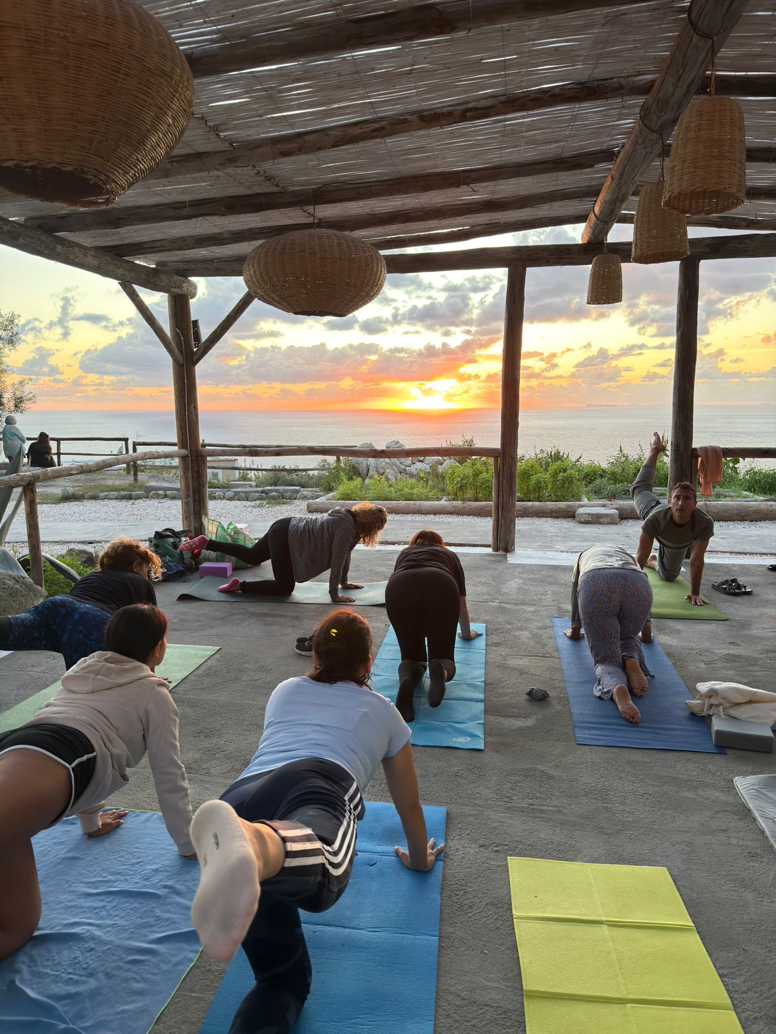 Un groupe de personnes pratiquant le yoga sur des tapis, sous une structure en bois, avec vue sur le coucher de soleil sur la mer.