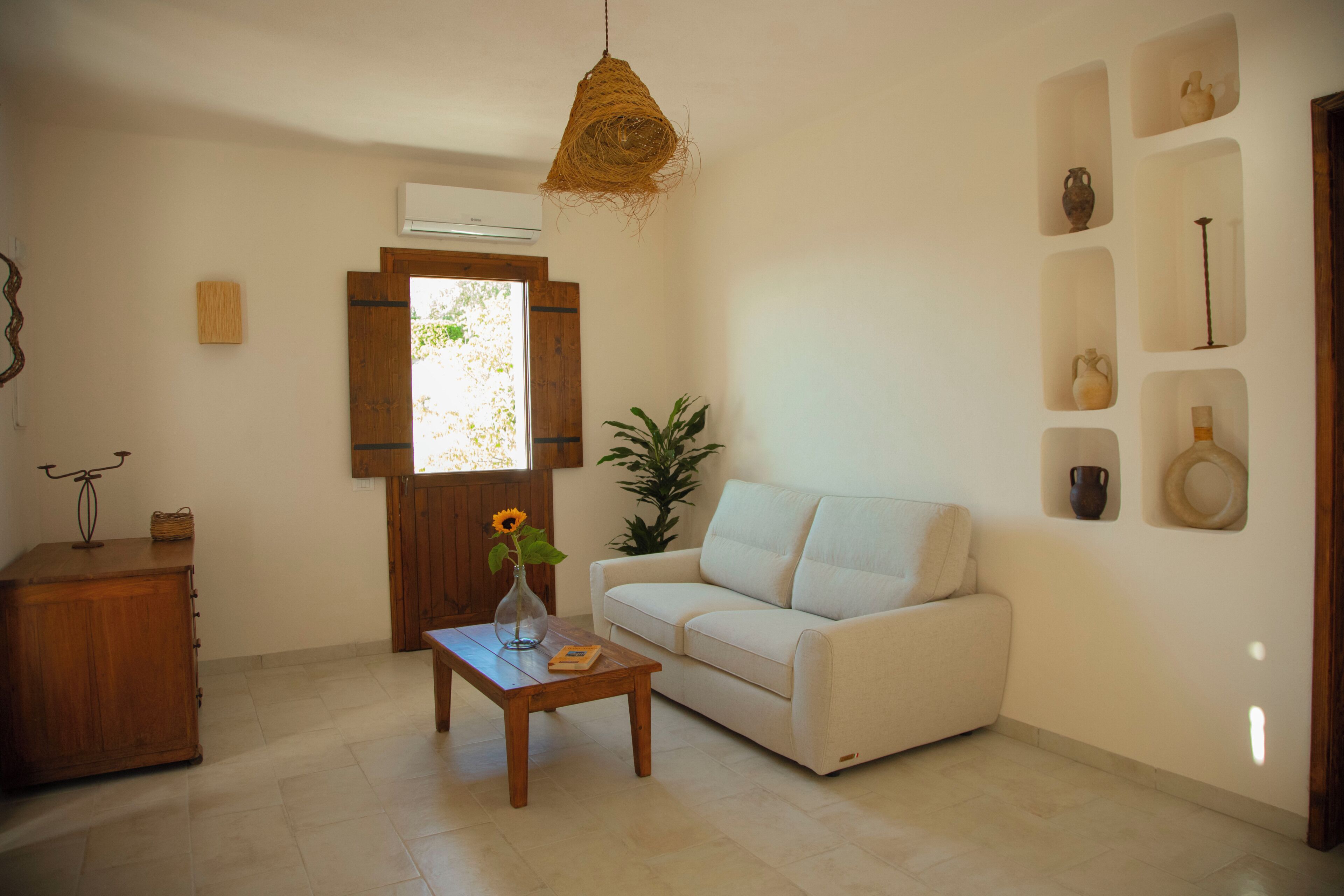A living room with a sofa, a wooden coffee table with a vase, a hanging light fixture, and a window with shutters.