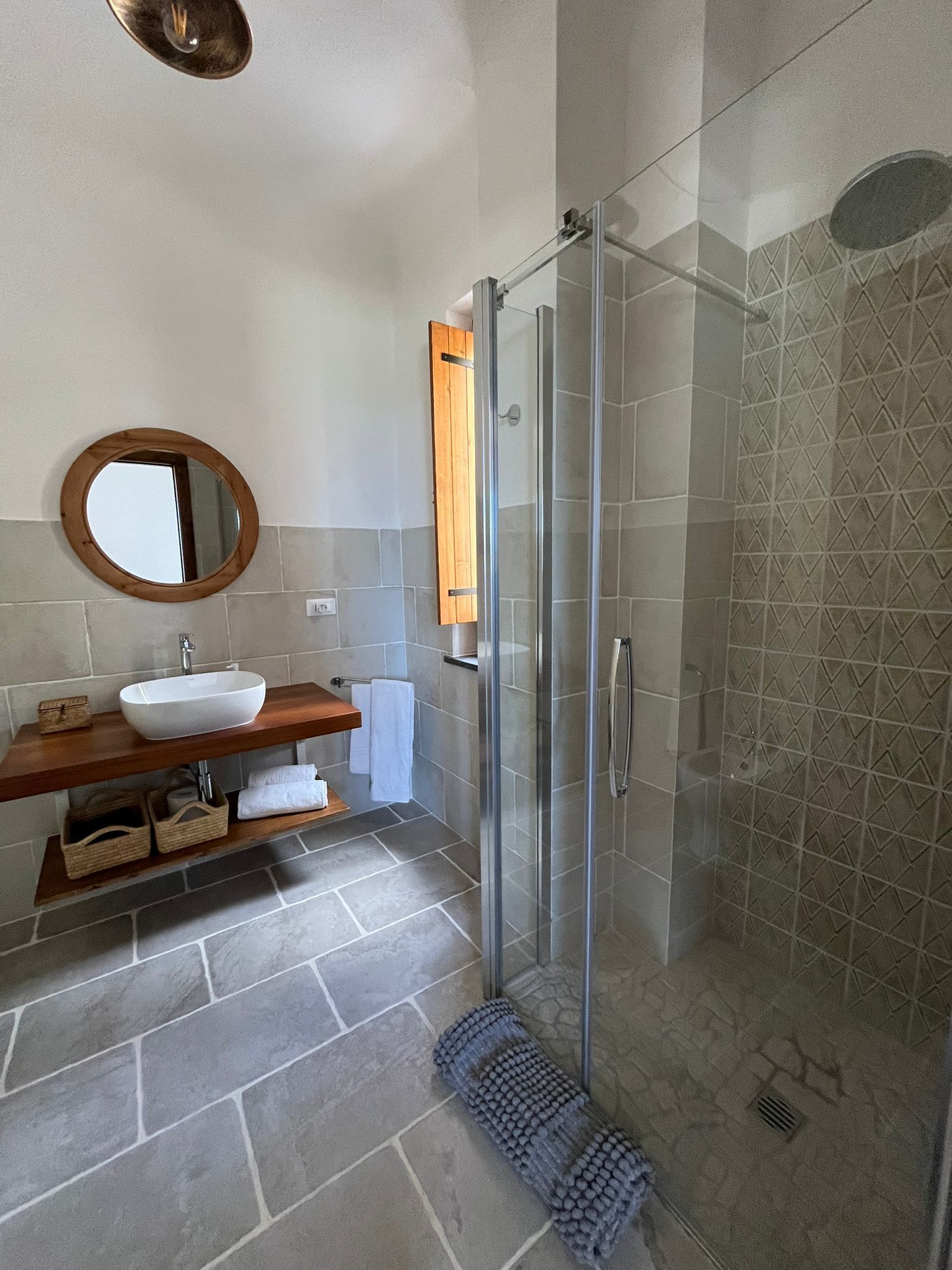 A bathroom with a glass-enclosed shower, a wooden vanity, a round mirror, and a white sink.