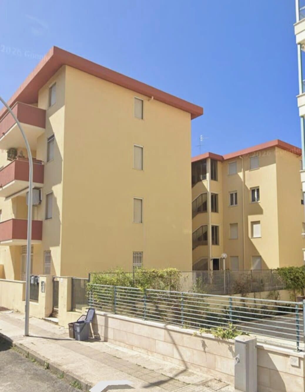 Um edifício residencial com vários andares, varandas, calçada pavimentada e cerca de metal.