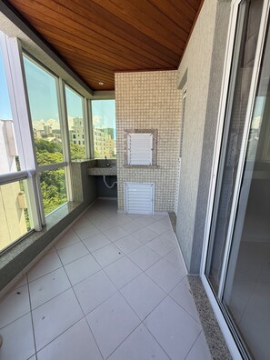 Un balcón con suelo de baldosas, techo de madera y vista a edificios y árboles.