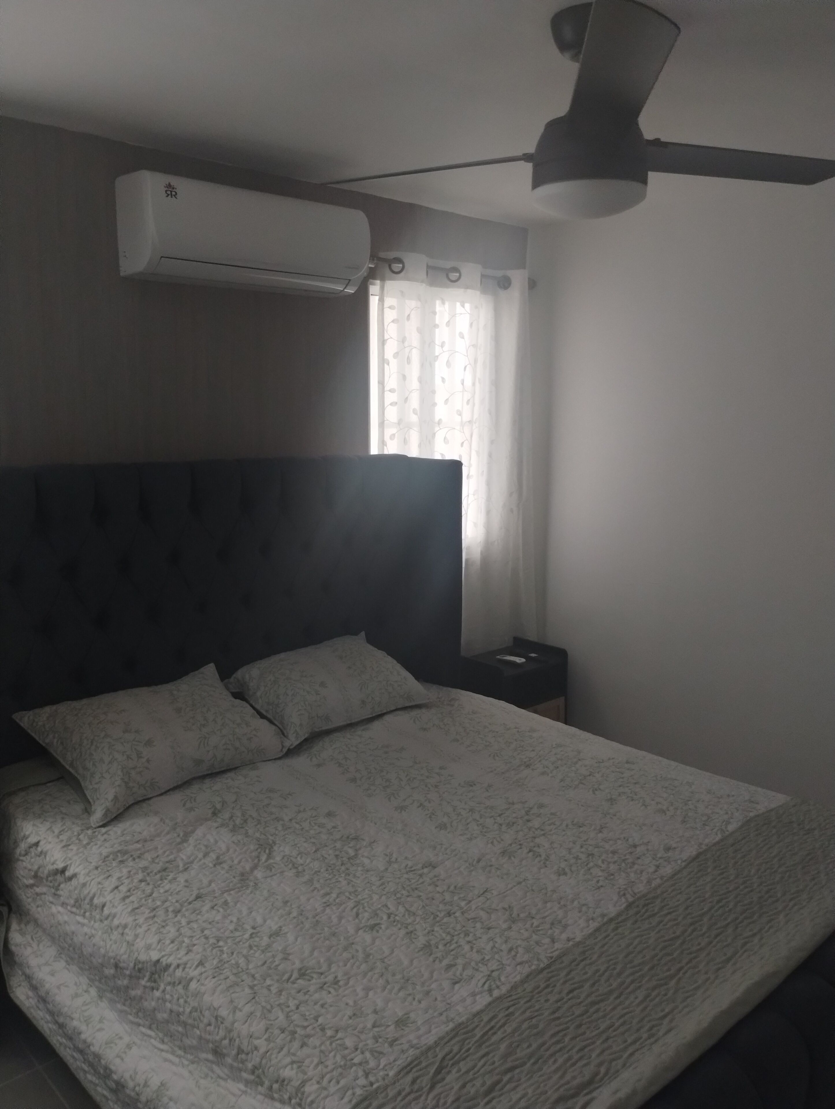 Een slaapkamer met een bed, nachtkastje, airconditioning en plafondventilator.