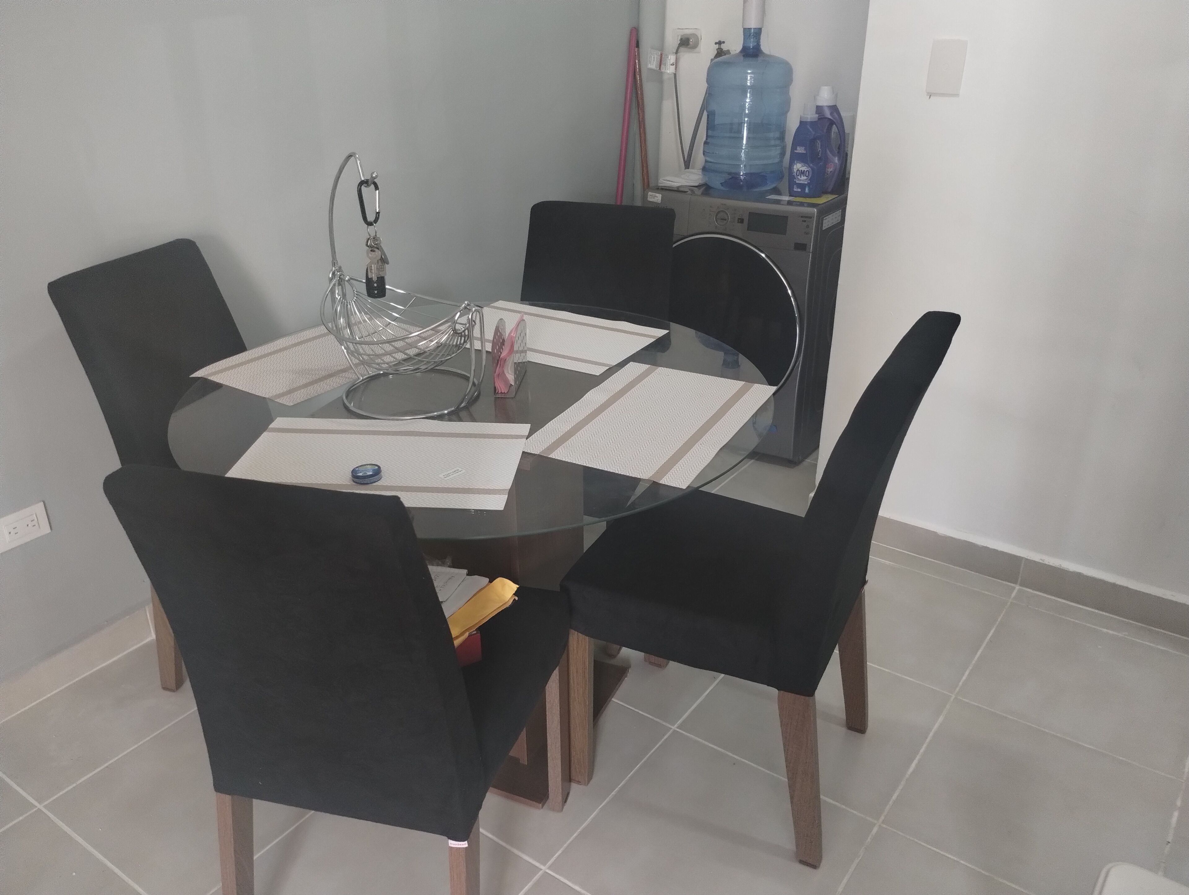 Een ronde glazen tafel met zwarte stoelen, een waterdispenser en een kleine verwarming aan de muur.