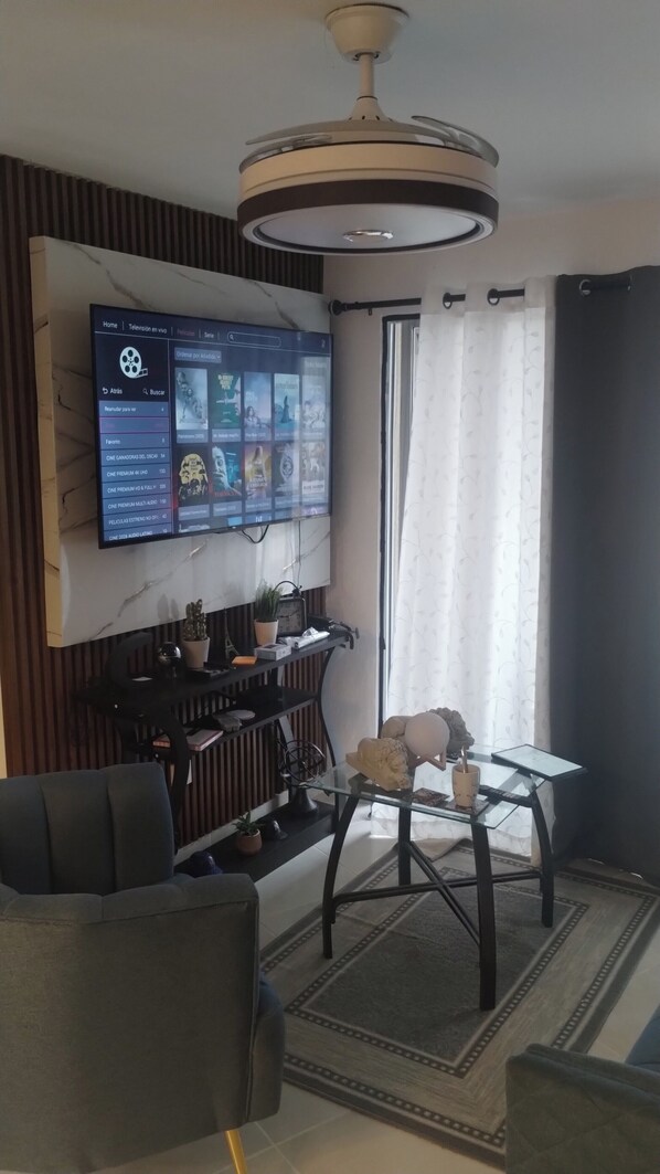 A living room with a flat-screen TV mounted on the wall, a glass coffee table, a sofa, and a small decorative table with a clock and plants.