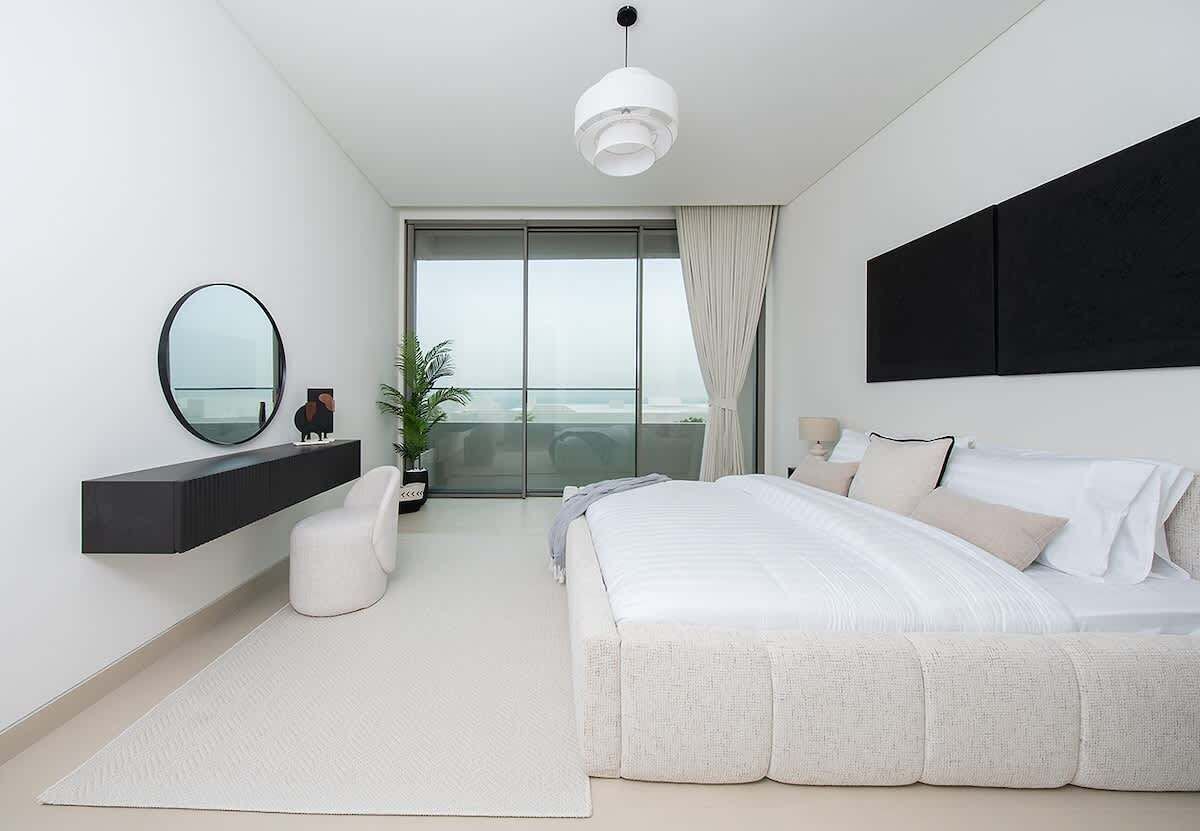 A modern bedroom with a large bed, a floating vanity, a round mirror, and a view of the outdoors.