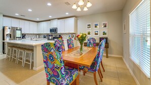 A modern kitchen with white cabinets, a central island, bar stools, colorful chairs, and a dining table with a vase of flowers. - Nature, Disney & More! Family Home in Groveland (Clermont)