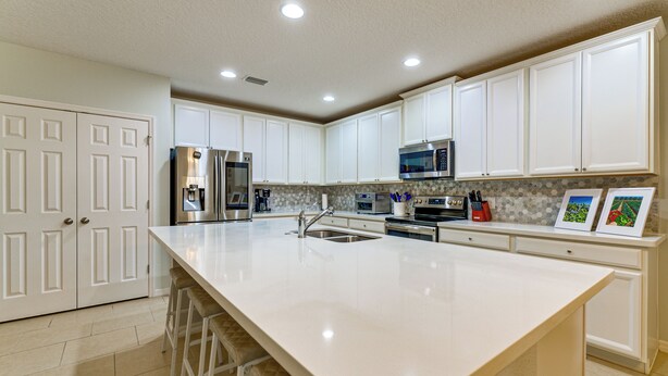 A kitchen with white cabinets, stainless steel appliances, a large island with stools, and a tiled backsplash. - Nature, Disney & More! Family Home in Groveland (Clermont)