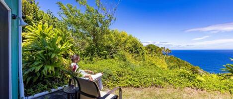 Una persona descansando en una silla junto a una ventana con vistas a un exuberante jardín y al océano.