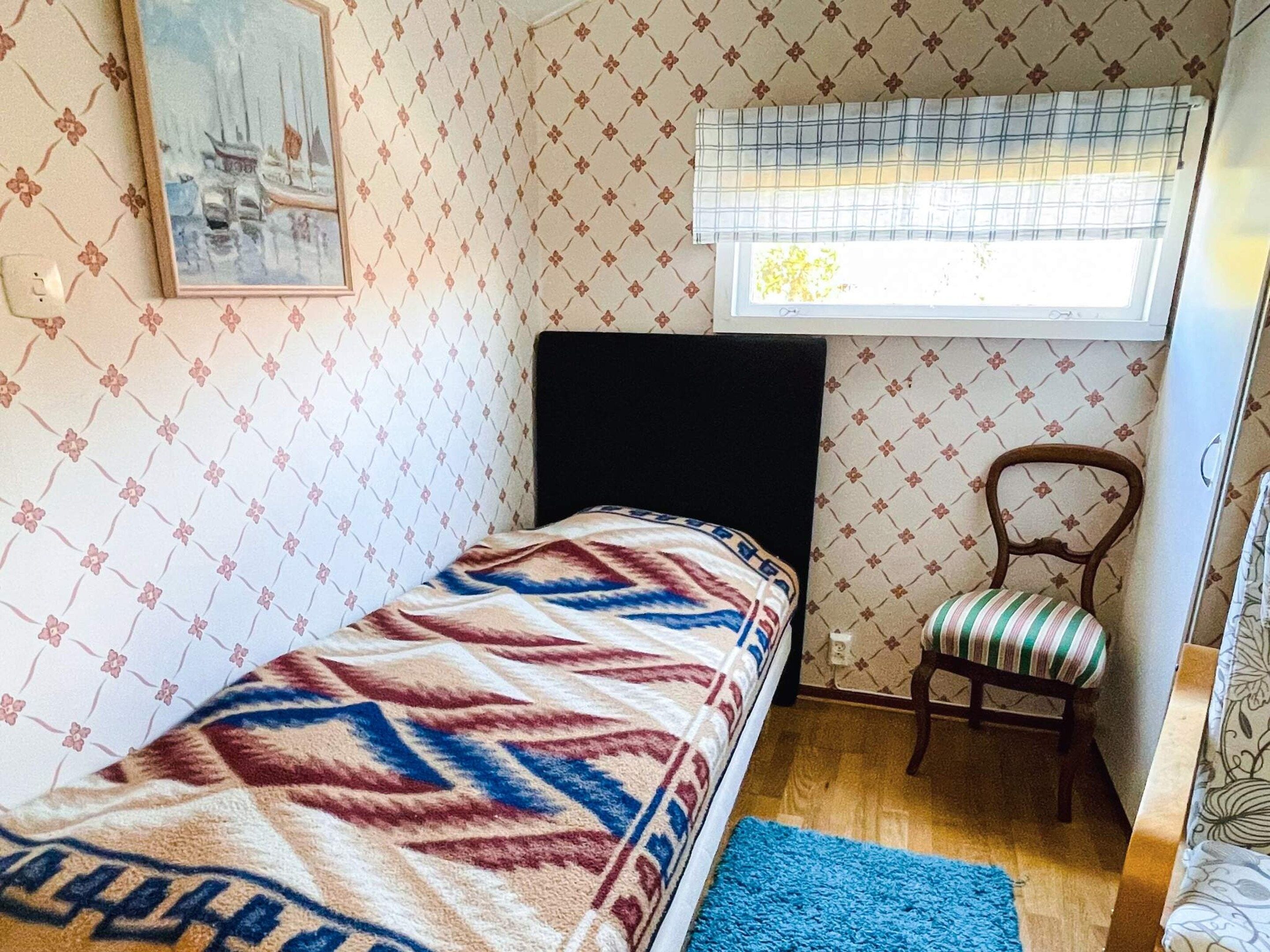 A small, single-bed room with a patterned wallpaper, a window with a blind, a chair, and a blue rug.