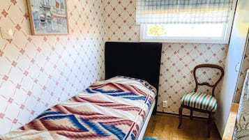 A small, single-bed room with a patterned wallpaper, a window with a blind, a chair, and a blue rug.