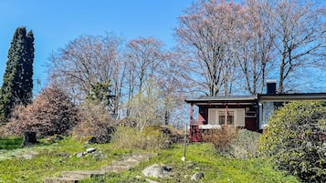Uma pequena casa com fachada vermelha, rodeada por um jardim com diversas plantas e árvores, algumas delas com flores cor-de-rosa.