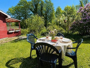 An outdoor dining area with a table set for four, surrounded by chairs, in a garden with a red building in the background. - 5 Person Holiday Home in Ronneby (Ronneby)