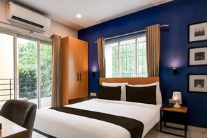 Una habitación de hotel con cama, escritorio, silla, mesita de noche con lámpara y ventana con cortinas.
