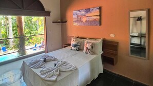 A bedroom with a bed, a ceiling fan, a window with a view of greenery, and a painting on the wall. - Mambuca Village (Ubatuba)