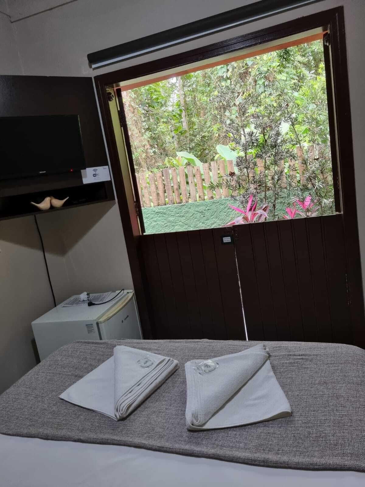 A room with a bed, a TV mounted on the wall, a small white appliance, and a window with a view of greenery.