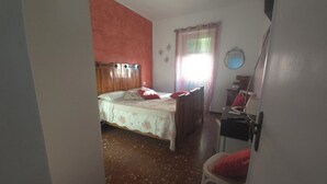 Quarto com cama, uma cadeira, escrivaninha, espelho e janela com cortinas.